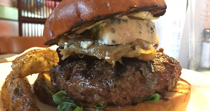 A tall grass-fed burger on a brioche bun topped with cheese and an onion ring.