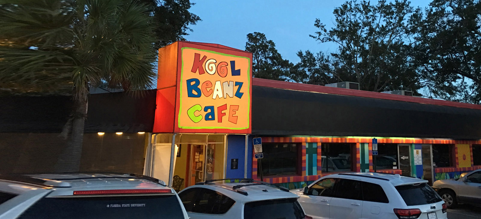 Kool Beanz Cafe at dusk with the sign lit up and cars parked in the lot out front.