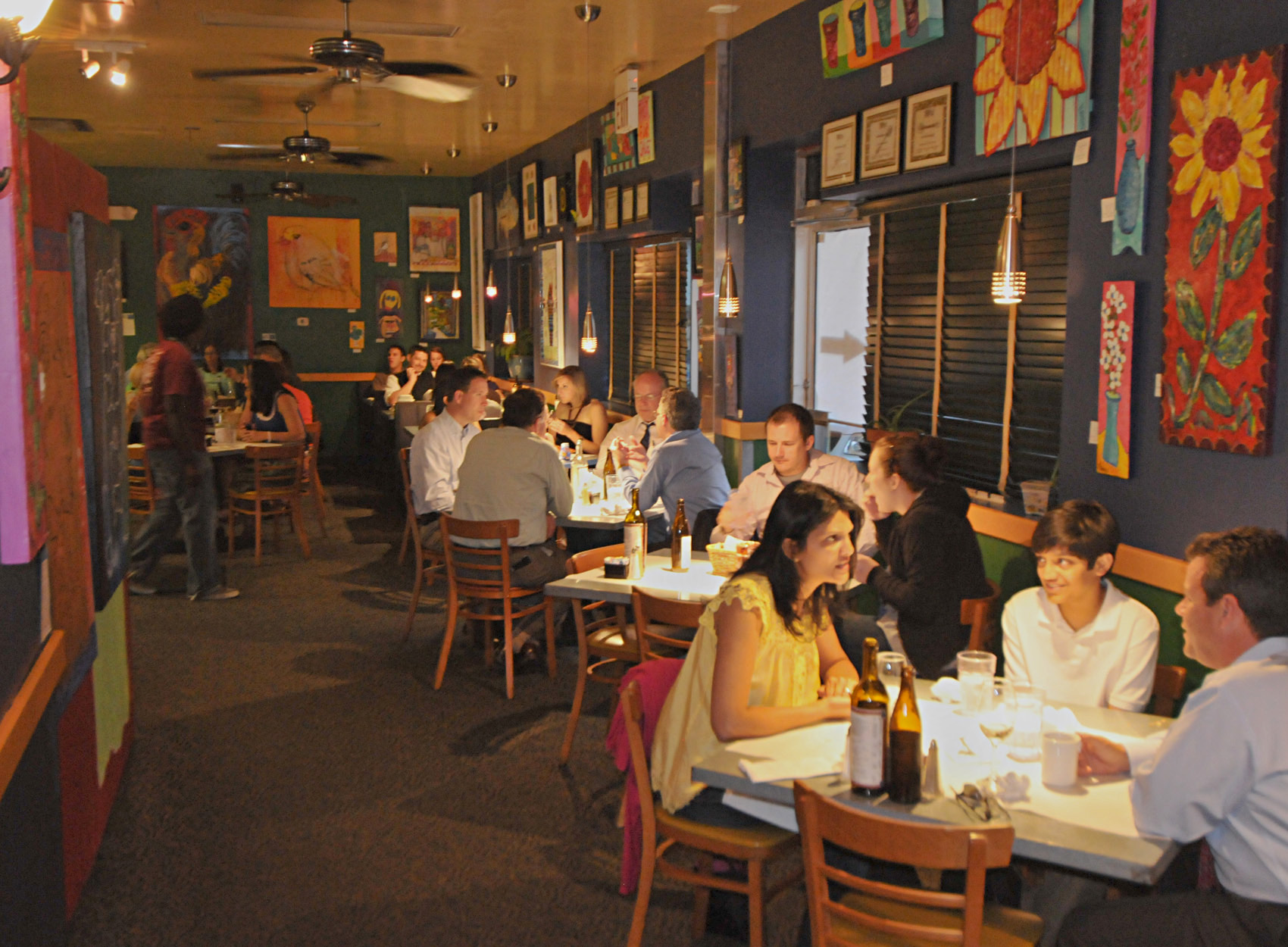 The Kool Beanz Cafe dining room full of guests at dinner, with colourful walls, hanging lamps and paintings above the tables.