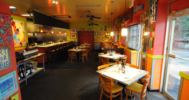 The main dining room at Kool Beanz Cafe, with painted orange and yellow walls, art hung close together, and the open kitchen along one side.