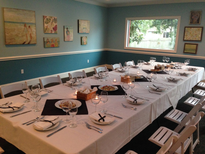 A long table in The Room at Kool Beanz set with white linen, wine glasses and dark napkins, with framed artwork on the blue-grey walls.